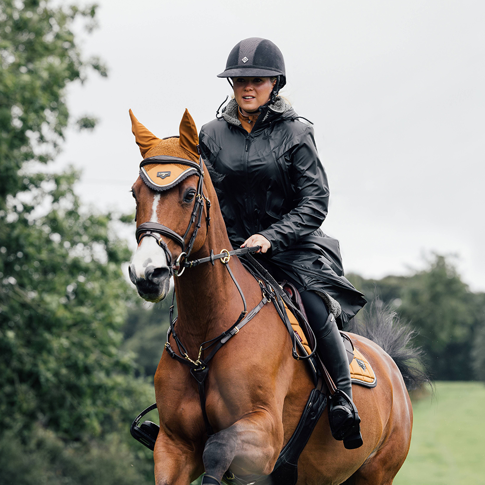 Woman riding a bay horse wearing the LeMieux All Elements Waterproof Horse Riding Coat in Black